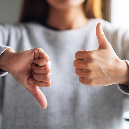 Person in grauem Pullover zeigt gleichzeitig Daumen hoch und Daumen runter, symbolisiert gemischtes Feedback oder Entscheidungsdilemma