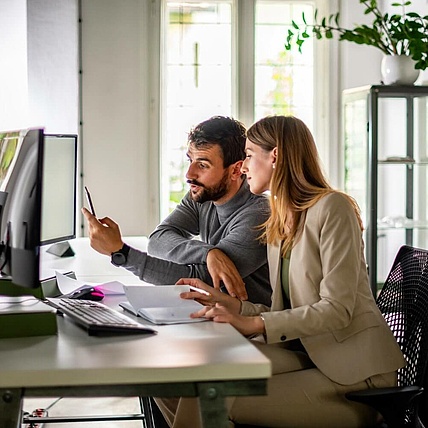 Ein Mann und eine Frau arbeiten gemeinsam an einem Schreibtisch, blicken auf einen Computerbildschirm und besprechen Dokumente.