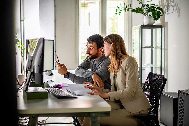 Ein Mann und eine Frau arbeiten gemeinsam an einem Schreibtisch, blicken auf einen Computerbildschirm und besprechen Dokumente.