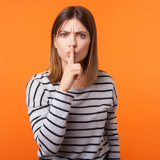 Junge Frau in gestreiftem Shirt vor orangefarbenem Hintergrund hält den Finger an die Lippen und signalisiert „Ruhe“